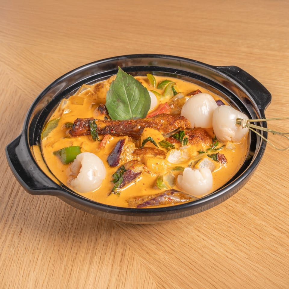 A black bowl of red curry with duck, lychees, and basil on a wooden table.