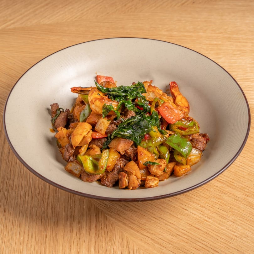 A Chinese dish with shrimp, beef, chicken, bell peppers, and basil in a light brown bowl.