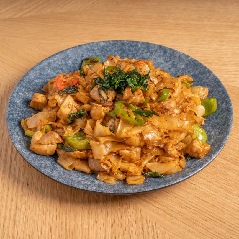 A plate of Chinese stir-fried noodles with chicken, green peppers, and basil on a wooden table.